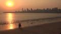 Three Friends Walk Along The Beach Towards The Skyline Of Mumbai At Sunset