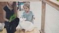 Young Beautiful Mother With Little Daughter Looking At Rabbits In Cage, Feeding