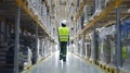 Female Warehouse Worker Wearing Helmet Checks Walks In Storehouse, Storeroom